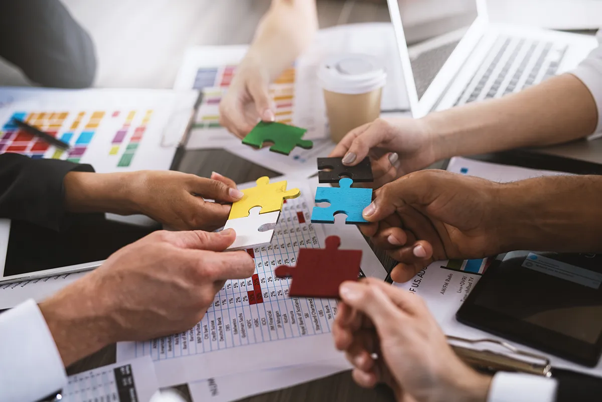 Teamarbeit mit Puzzleteilen im Büro Mehrere Hände von Geschäftspersonen halten verschiedenfarbige Puzzleteile über einem Schreibtisch, um Zusammenarbeit und Teamwork zu symbolisieren.